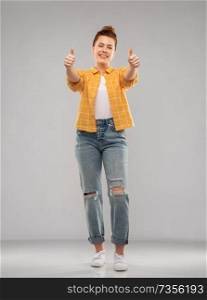 gesture and people concept - smiling red haired teenage girl in checkered shirt and torn jeans showing thumbs up over grey background. happy red haired teenage girl showing thumbs up