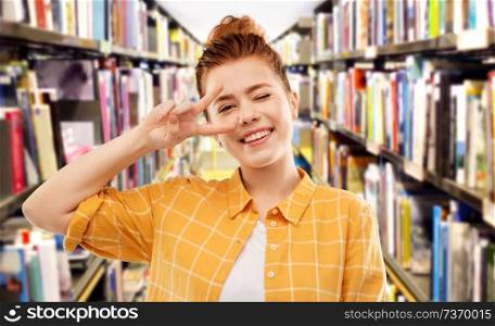 gesture and people concept - smiling red haired teenage student girl in checkered shirt showing peace hand sign over library background. red haired student girl showing peace at library