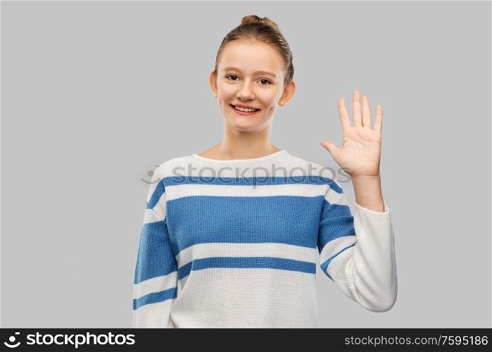 gesture and people concept - smiling teenage girl in pullover waving hand over grey background. smiling teenage girl in pullover waving hand