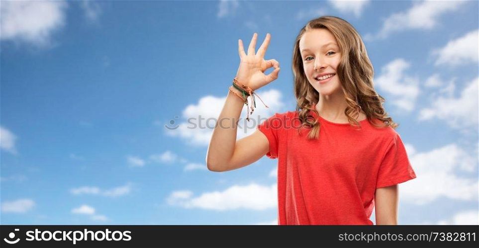 gesture and people concept - smiling teenage girl with long hair in red t-shirt showing ok hand sign over blue sky and clouds background. smiling teenage girl in red t-shirt showing ok
