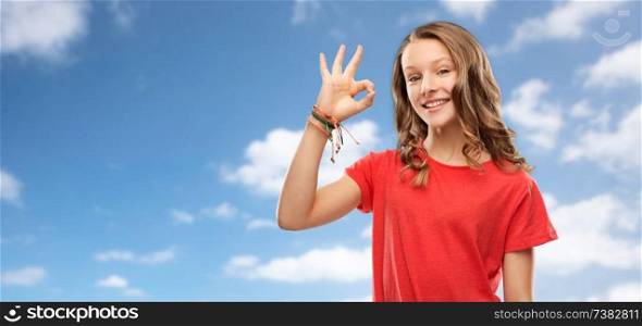 gesture and people concept - smiling teenage girl with long hair in red t-shirt showing ok hand sign over blue sky and clouds background. smiling teenage girl in red t-shirt showing ok