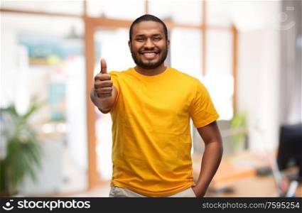 gesture and people concept - smiling young african american man in yellow t-shirt showing thumbs up over office background. smiling african american man showing thumbs up