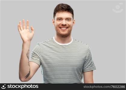 gesture and people concept - smiling young man in striped t-shirt waving hand over grey background. smiling young man in striped t-shirt waving hand