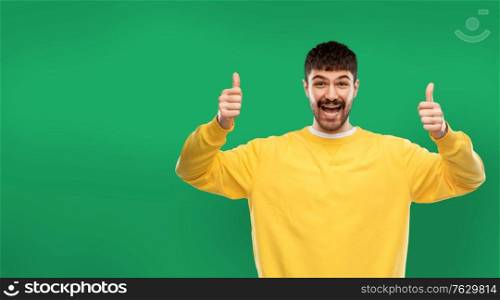 gesture and people concept - smiling young man in yellow sweatshirt showing thumbs up over green background. happy man showing thumbs up over green background