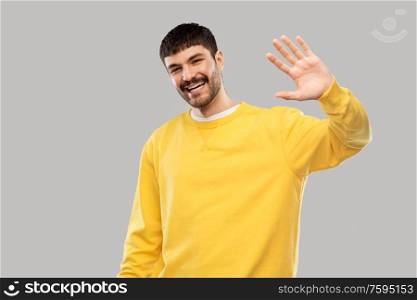 gesture and people concept - smiling young man in yellow sweatshirt waving hand over grey background. smiling young man in yellow sweatshirt waving hand
