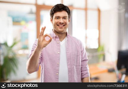 gesture and people concept - smiling young man showing ok hand sign over office room background. smiling young man showing ok hand sign over office