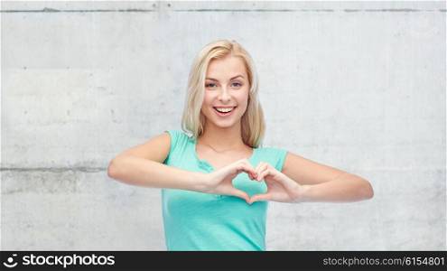 gesture and people concept - smiling young woman or teenage girl showing heart shape made of fingers over gray concrete wall background. happy woman or teen girl showing heart shape sigh