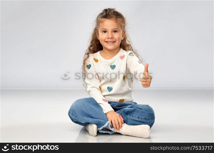 gesture, childhood and people concept - beautiful smiling girl sitting on floor and showing thumbs up over grey background. little girl sitting on floor and showing thumbs up