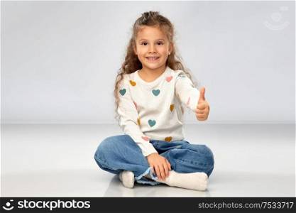 gesture, childhood and people concept - beautiful smiling girl sitting on floor and showing thumbs up over grey background. little girl sitting on floor and showing thumbs up