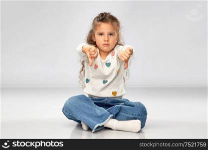 gesture, childhood and people concept - sad girl sitting on floor and showing thumbs down over grey background. sad girl sitting on floor and showing thumbs down