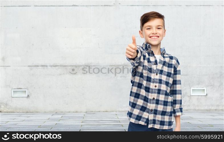 gesture, childhood, gender, fashion and people concept - smiling boy in checkered shirt and jeans showing thumbs up over urban street background