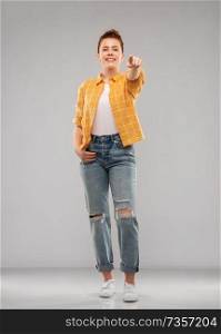 gesture, choice and people concept - smiling red haired teenage girl in checkered shirt and torn jeans pointing finger to you over grey background. red haired teenage girl pointing finger to you