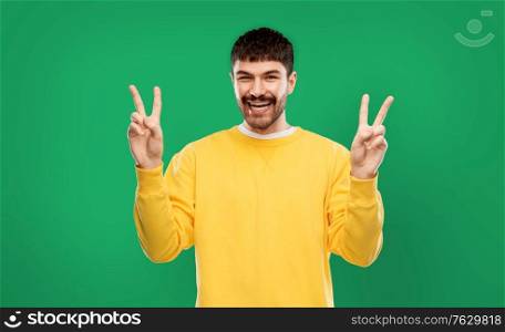 gesture, count and people concept - happy smiling young man in yellow sweatshirt showing peace hand sign over emerald green background. young man showing peace over grey background