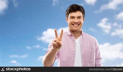 gesture, count and people concept - smiling young man showing two fingers or peace hand sign over blue sky and clouds background. man showing two fingers or peace sign over sky