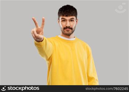 gesture, count and people concept - young man in yellow sweatshirt showing peace hand sign over grey background. young man showing peace over grey background