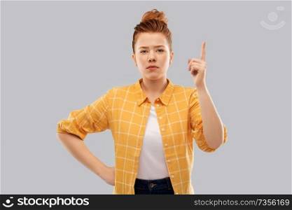 gesture, counting and people concept - serious red haired teenage girl in checkered shirt pointing finger up over grey background. red haired teenage girl pointing finger up