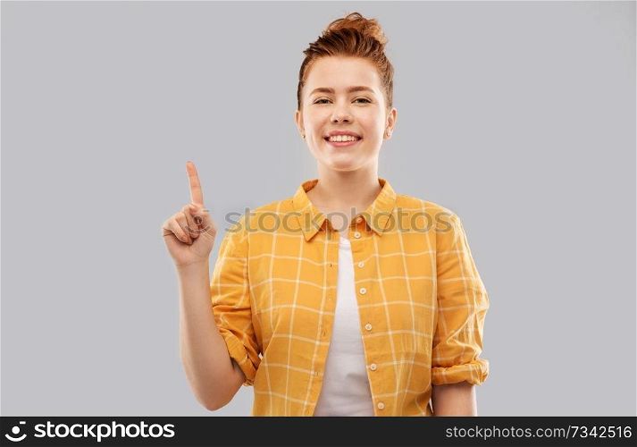 gesture, counting and people concept - smiling red haired teenage girl in checkered shirt showing one finger or pointing up over grey background. red haired teenage girl showing one finger