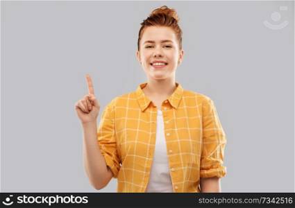 gesture, counting and people concept - smiling red haired teenage girl in checkered shirt showing one finger or pointing up over grey background. red haired teenage girl showing one finger