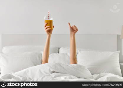 gesture, drink and morning concept - hands of young woman lying in bed with cup of juice showing thumbs up at bedroom. hands of woman lying in bed with cup of juice