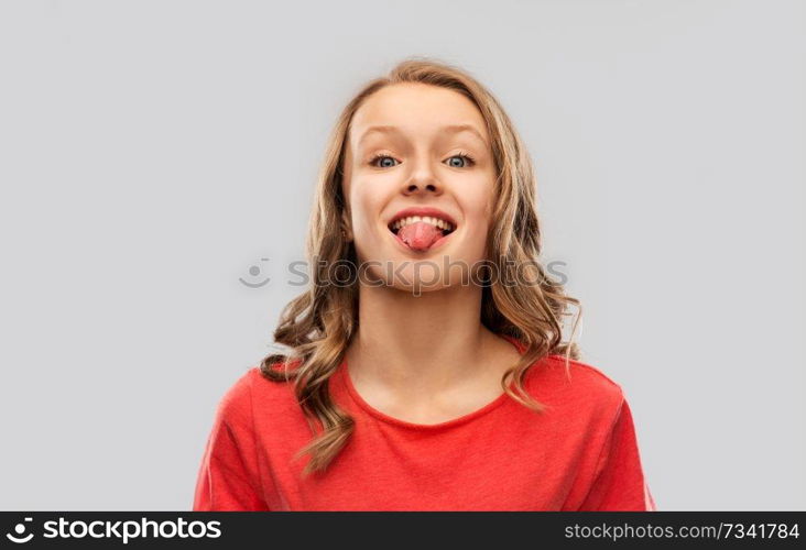 gesture, expression and people concept - funny teenage girl with long hair in red t-shirt showing tongue over grey background. funny teenage girl in red t-shirt showing tongue