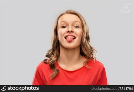 gesture, expression and people concept - funny teenage girl with long hair in red t-shirt showing tongue over grey background. funny teenage girl in red t-shirt showing tongue