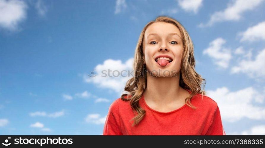gesture, expression and people concept - funny teenage girl with long hair in red t-shirt showing tongue over blue sky and clouds background. teenage girl in red t-shirt shows tongue over sky