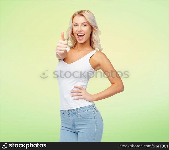 gesture, expressions and people concept - happy smiling young woman in white top and jeans showing thumbs up over green background. happy young woman showing thumbs up