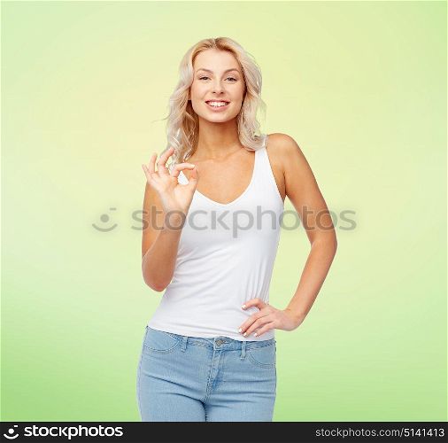 gesture, expressions and people concept - happy smiling young woman in white top and jeans showing ok hand sigh over green background. happy young woman showing ok hand sigh