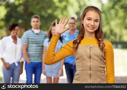 gesture, friendship and people concept - happy smiling young teenage girl in mustard yellow top waving hand over group of friends at park on background. happy teenage girl waving hand over friends