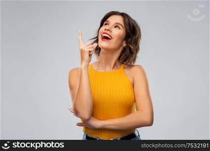 gesture, idea and people concept - happy smiling young woman in mustard yellow top pointing finger up over grey background. happy smiling young woman pointing finger up