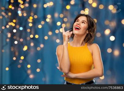 gesture, idea and people concept - happy smiling young woman in yellow top pointing finger up over festive lights on dark blue background. happy smiling young woman pointing finger up