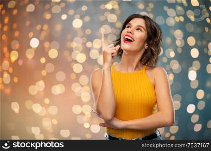 gesture, idea and people concept - happy smiling young woman in yellow top pointing finger up over festive lights background. happy smiling young woman pointing finger up