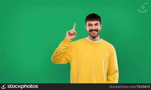 gesture, idea and people concept - smiling young man in yellow sweatshirt pointing one finger up over emerald green background. happy man pointing finger up over green