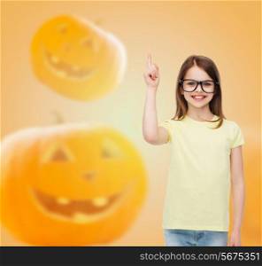 gesture, idea, education, holidays and people concept - smiling little girl in glasses pointing finger over halloween pumpkins background