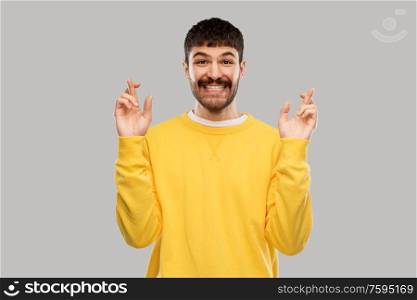 gesture, luck and people concept - smiling young man with crossed fingers in yellow sweatshirt over grey background. smiling man with crossed fingers