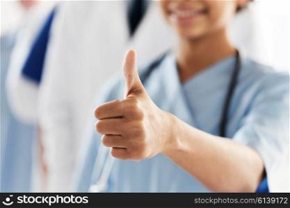 gesture, people, health care and medicine concept - close up of happy african female doctor or nurse showing thumbs up hand sign over group of medics meeting at hospital