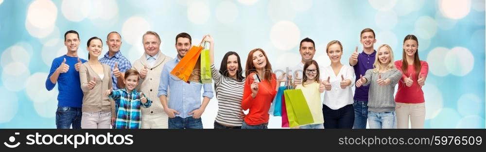 gesture, sale and people concept - group of smiling men, women and kids showing thumbs up and holding shopping bags with credit card over blue holidays lights background
