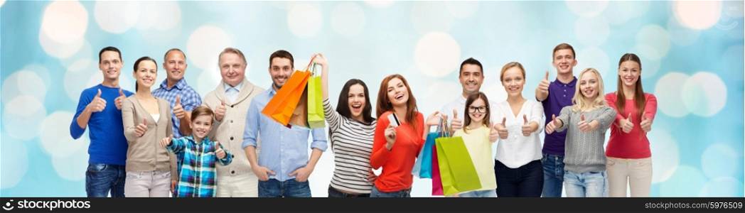 gesture, sale and people concept - group of smiling men, women and kids showing thumbs up and holding shopping bags with credit card over blue holidays lights background