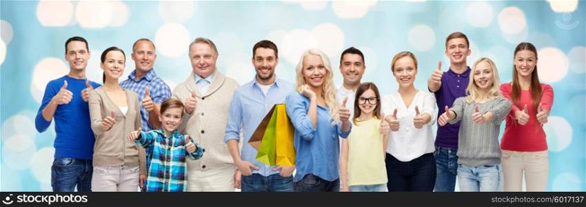 gesture, sale and people concept - group of smiling men, women and kids showing thumbs up and holding shopping bags over blue holidays lights background