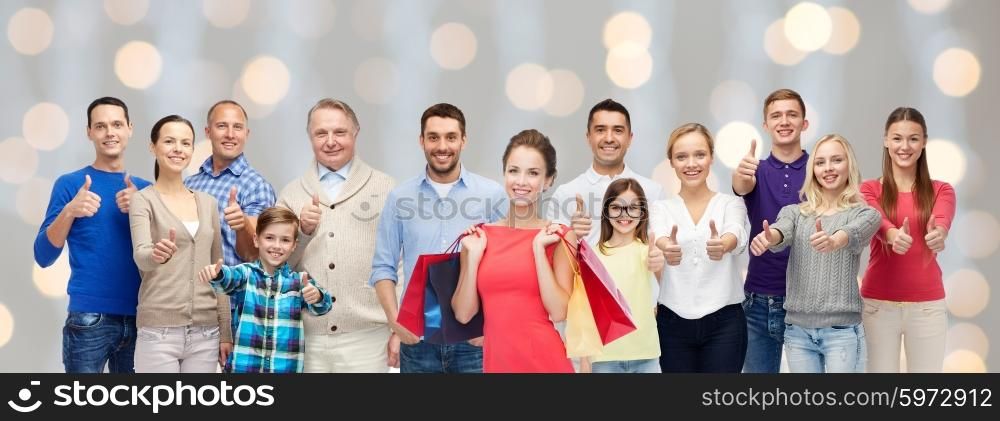 gesture, sale, shopping and people concept - group of smiling men, women and kids showing thumbs up and holding shopping bags over holidays lights background