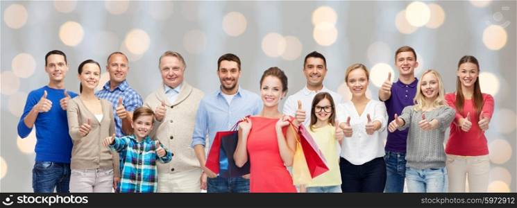 gesture, sale, shopping and people concept - group of smiling men, women and kids showing thumbs up and holding shopping bags over holidays lights background