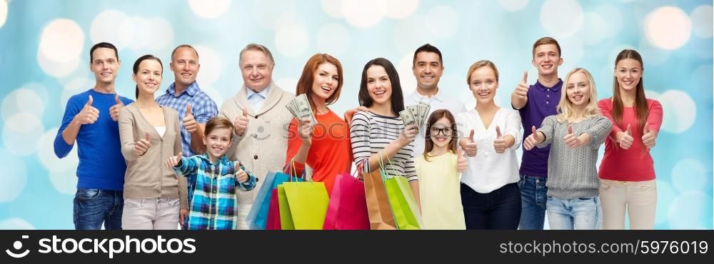 gesture, sale, shopping and people concept - group of smiling men, women and kids showing thumbs up and holding shopping bags with money over blue holidays lights background