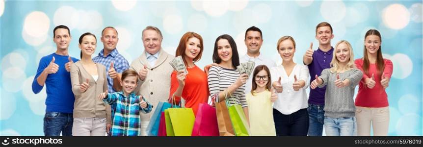 gesture, sale, shopping and people concept - group of smiling men, women and kids showing thumbs up and holding shopping bags with money over blue holidays lights background