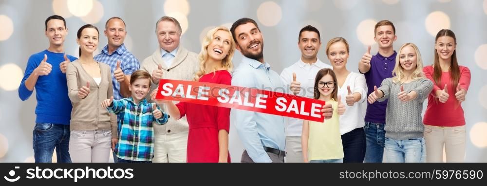 gesture, sale, shopping and people concept - group of smiling men, women and kids showing thumbs up and holding red sale sign or banner over holidays lights background
