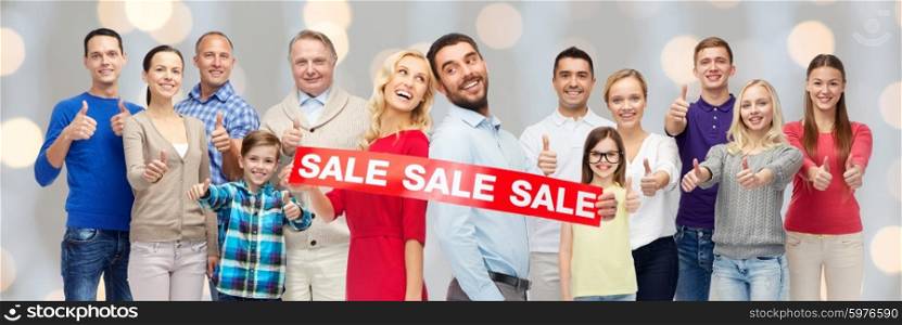 gesture, sale, shopping and people concept - group of smiling men, women and kids showing thumbs up and holding red sale sign or banner over holidays lights background