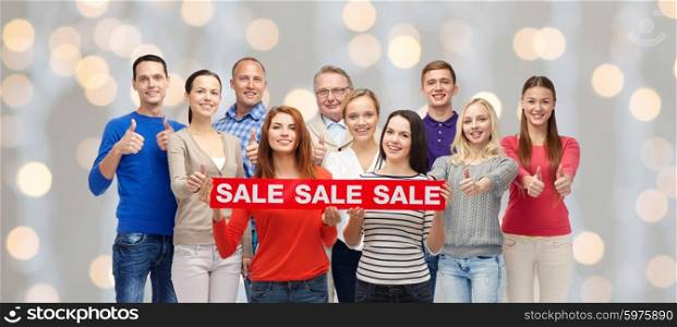 gesture, shopping and people concept - group of smiling men and women showing thumbs up and holding red sale sign or banner over holidays lights background