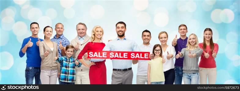 gesture, shopping and people concept - group of smiling men, women and kids showing thumbs up and holding red sale sign or banner over blue holidays lights background