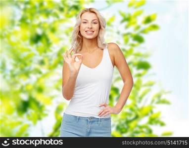 gesture, summer and people concept - happy smiling young woman in white top and jeans showing ok hand sigh over green natural background. happy young woman showing ok hand sigh