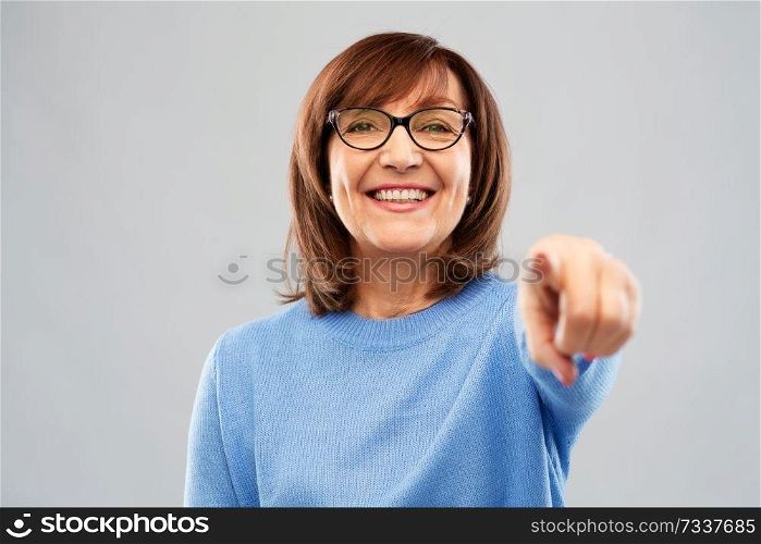 gesture, vision and old people concept - portrait of smiling senior woman in glasses pointing finger to you over grey background. senior woman in glasses pointing finger to you