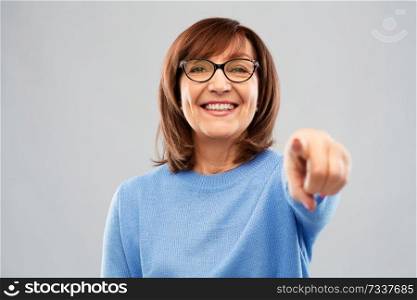 gesture, vision and old people concept - portrait of smiling senior woman in glasses pointing finger to you over grey background. senior woman in glasses pointing finger to you
